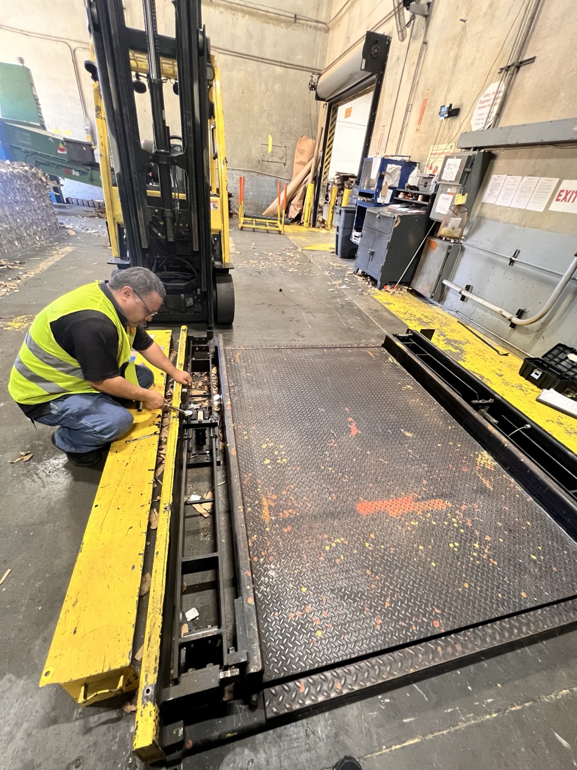A scale technician in a hi-visibility vest kneeling to service the weighing platform of a forklift scale on a plant floor.